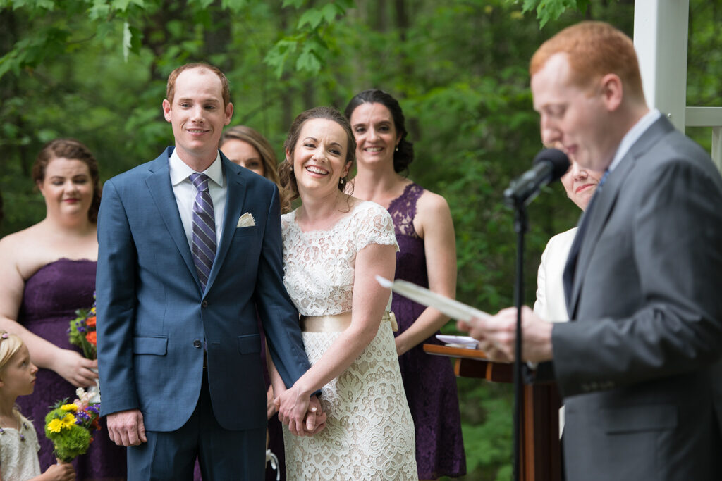 marrying couple smile as they listen to a reading during their outdoor ceremony in New Hampshire